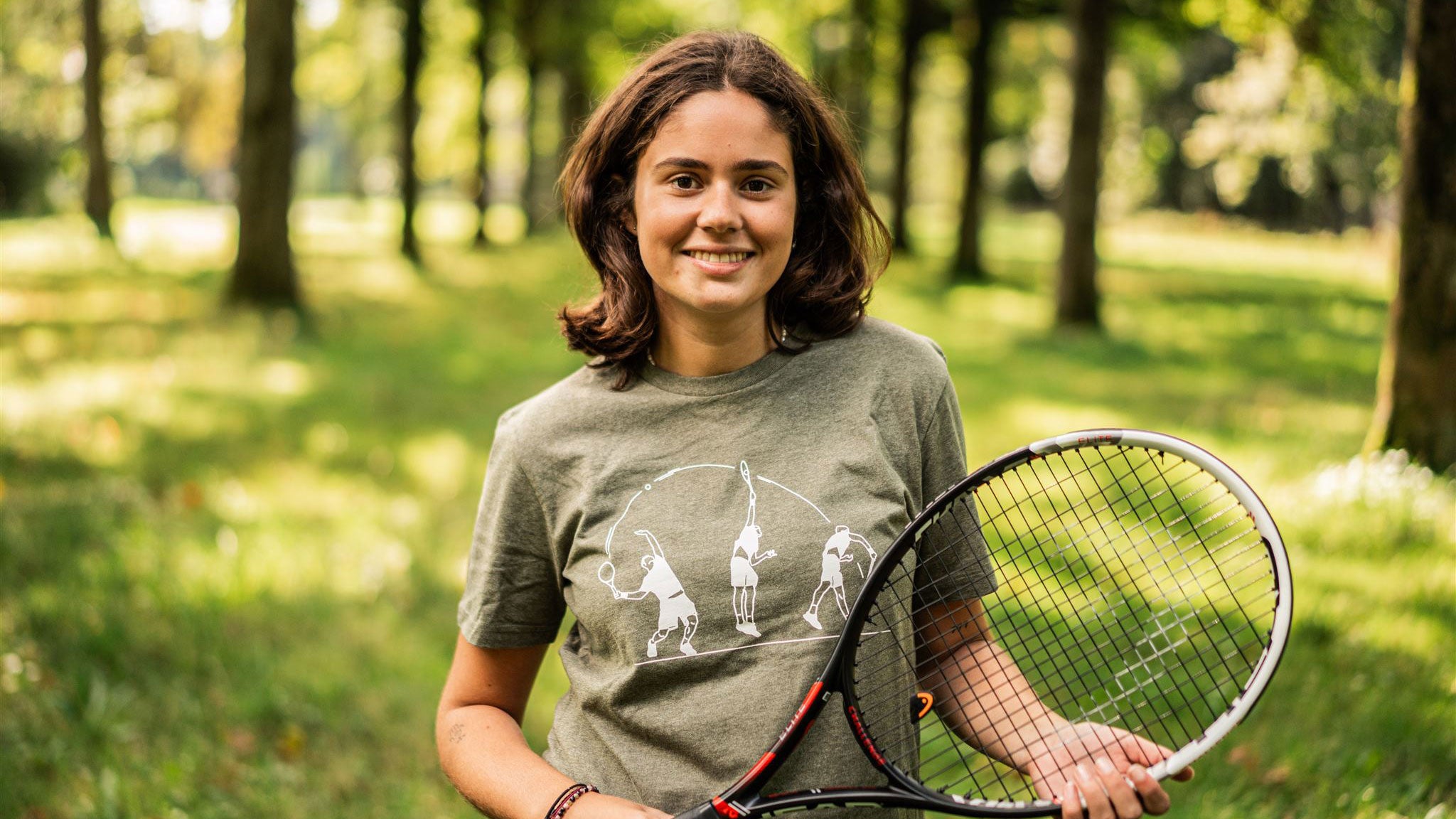 Woman with a tennis racket wearing a Khaki T-shirt with an illustration of tennis on it.